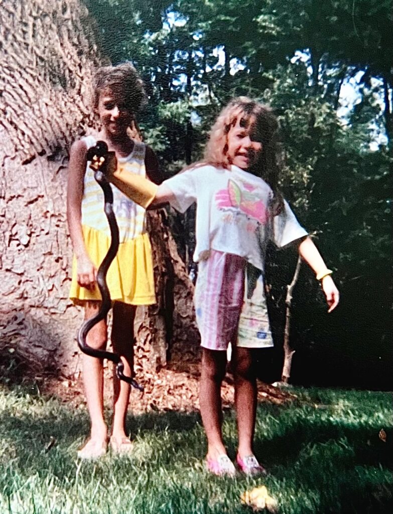 Two sisters pose, one in a yellow dress and the other in a white top and pink shorts with a yellow glove holding a black snake by its neck.